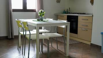 Un comedor con una mesa y sillas blancas, una planta pequeña en maceta y una cocina con armarios y electrodomésticos.