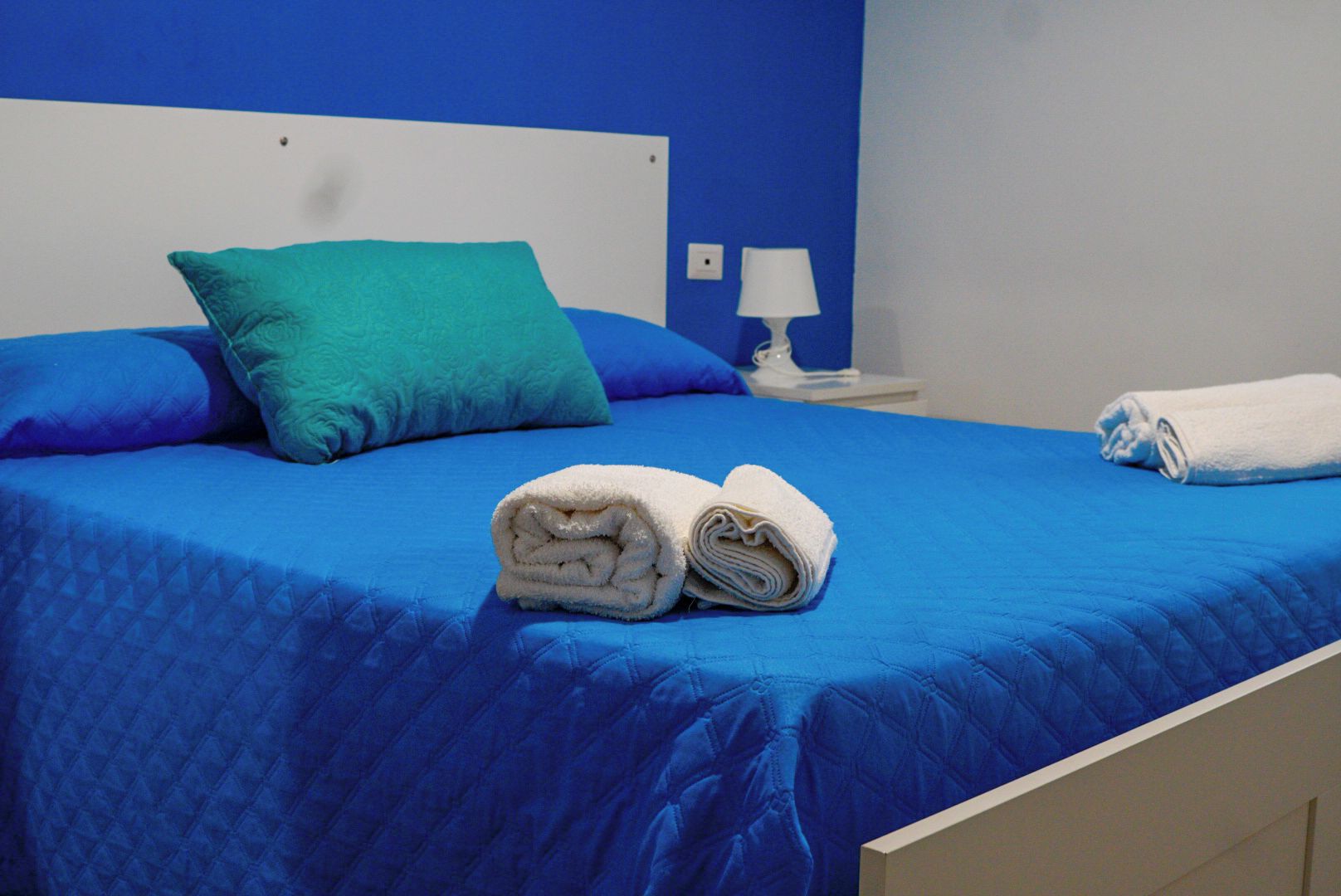 Ein Bett mit blauer Bettwäsche, einem türkisfarbenen Kopfkissen und zwei gerollten weißen Handtüchern.