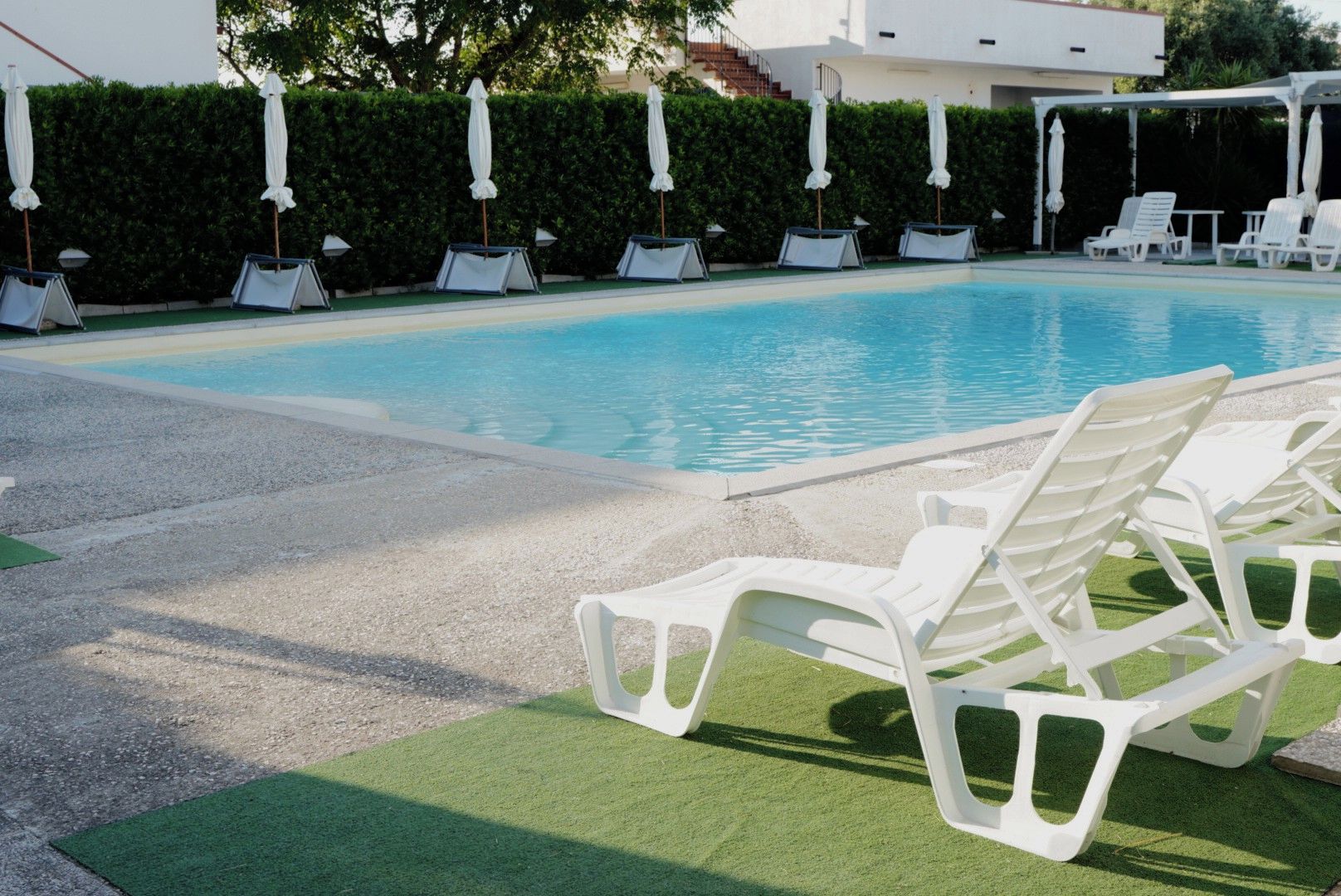 Piscina con sillas de descanso blancas y sombrillas, rodeada por un seto y un edificio.