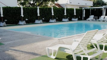 Piscina con sillas de descanso blancas y sombrillas, rodeada por un seto y un edificio.