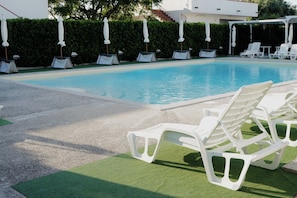 En swimmingpool med hvide liggestole og parasoller, omgivet af en hæk og en bygning.