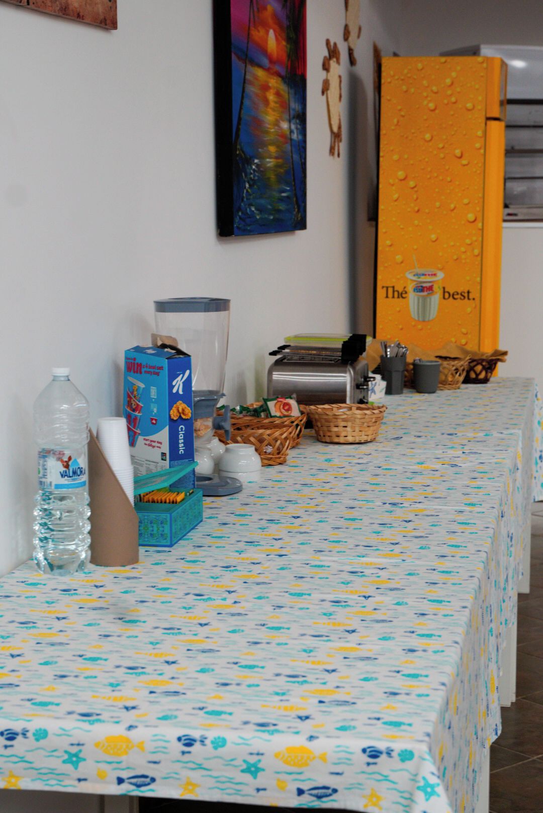Ein Tisch mit einer gemusterten Tischdecke, einer Wasserflasche, einer Milchkarton, einem Toaster und verschiedenen Körben.