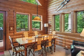 Ein Holzblockhaus-Interieur mit Esstisch, Stühlen und großen Fenstern, die einen Blick auf den Wald bieten.