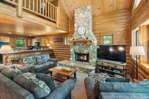 A log cabin interior with a stone fireplace, a television, and a kitchen area. - Cozy Log Cabin Retreat with Hot Tub (Boyne City)