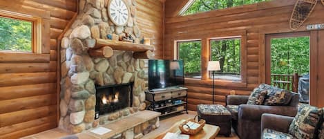 Interior de una cabaña de madera con chimenea de piedra, televisor y una zona de estar acogedora.