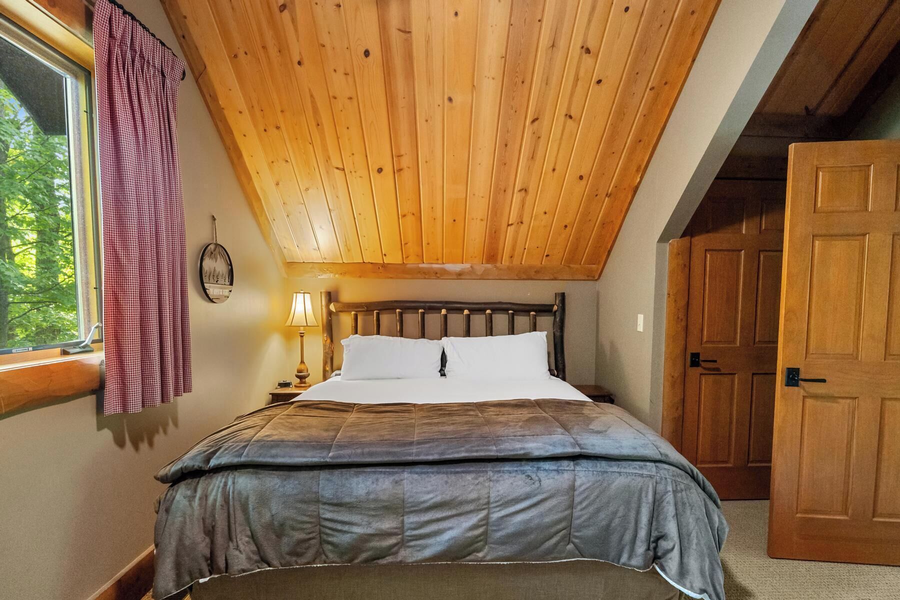 A bedroom with a wooden ceiling, a bed with a grey duvet, white pillows, a bedside lamp, and a window with a checkered curtain.