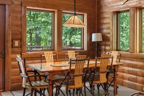 A wooden cabin interior with a dining table surrounded by chairs, large windows, and a hanging light fixture. - Cozy Log Cabin Retreat with Hot Tub (Boyne City)