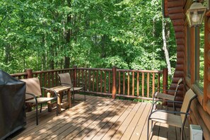 A wooden deck with outdoor furniture, surrounded by dense green foliage. - Cozy Log Cabin Retreat with Hot Tub (Boyne City)