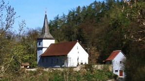 Uma igreja com um pináculo e um edifício branco com telhado vermelho, cercados por árvores e um muro de pedra.