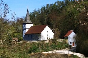 Una chiesa con un campanile e un edificio bianco con tetto rosso, circondati da alberi e un muro in pietra.