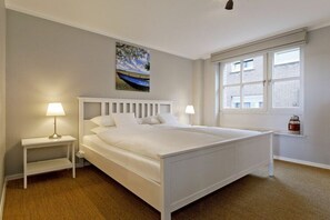 A bedroom with a large bed, two bedside tables with lamps, a window with blinds, and a painting on the wall. - Salmon fishermen in their cottage on the Rhine (Rees)