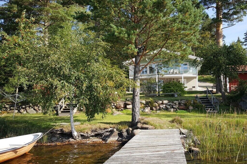 Un molo di legno conduce a una casa con un balcone che si affaccia su uno specchio d'acqua, circondata da alberi e un muro di pietra.