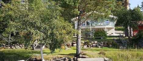 A wooden dock leading to a house with a balcony overlooking a body of water, surrounded by trees and a stone wall.