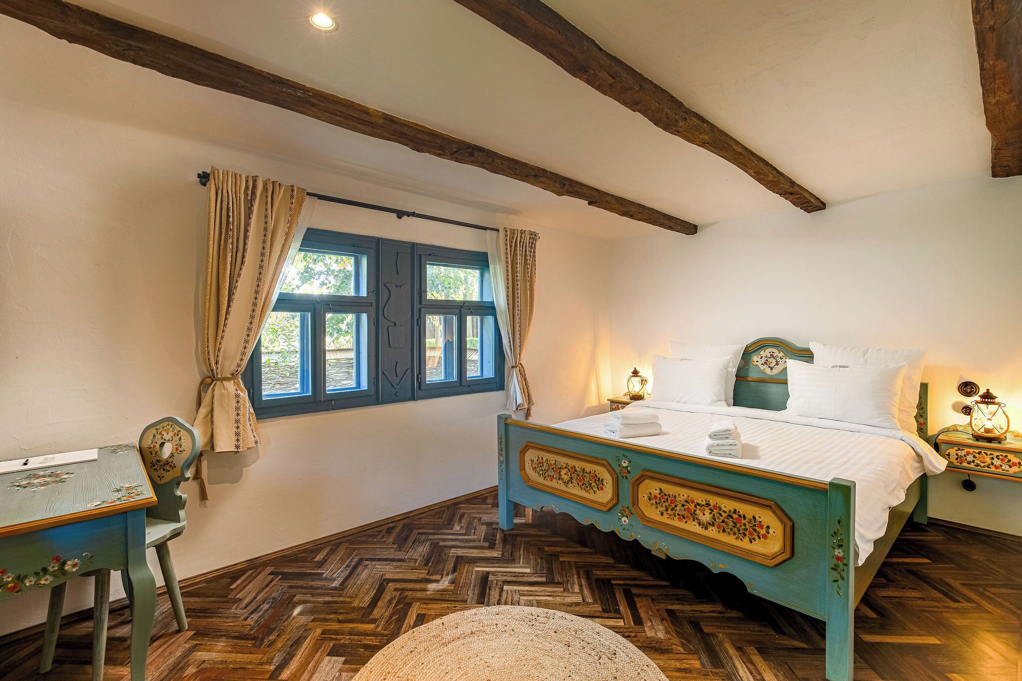 Un dormitorio con piso de madera tipo espiga, una cama con cabecera floral, un escritorio con silla y una ventana con cortinas.