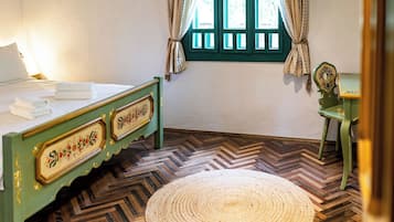 A room with a bed, a chair, a window with curtains, and a herringbone patterned floor.