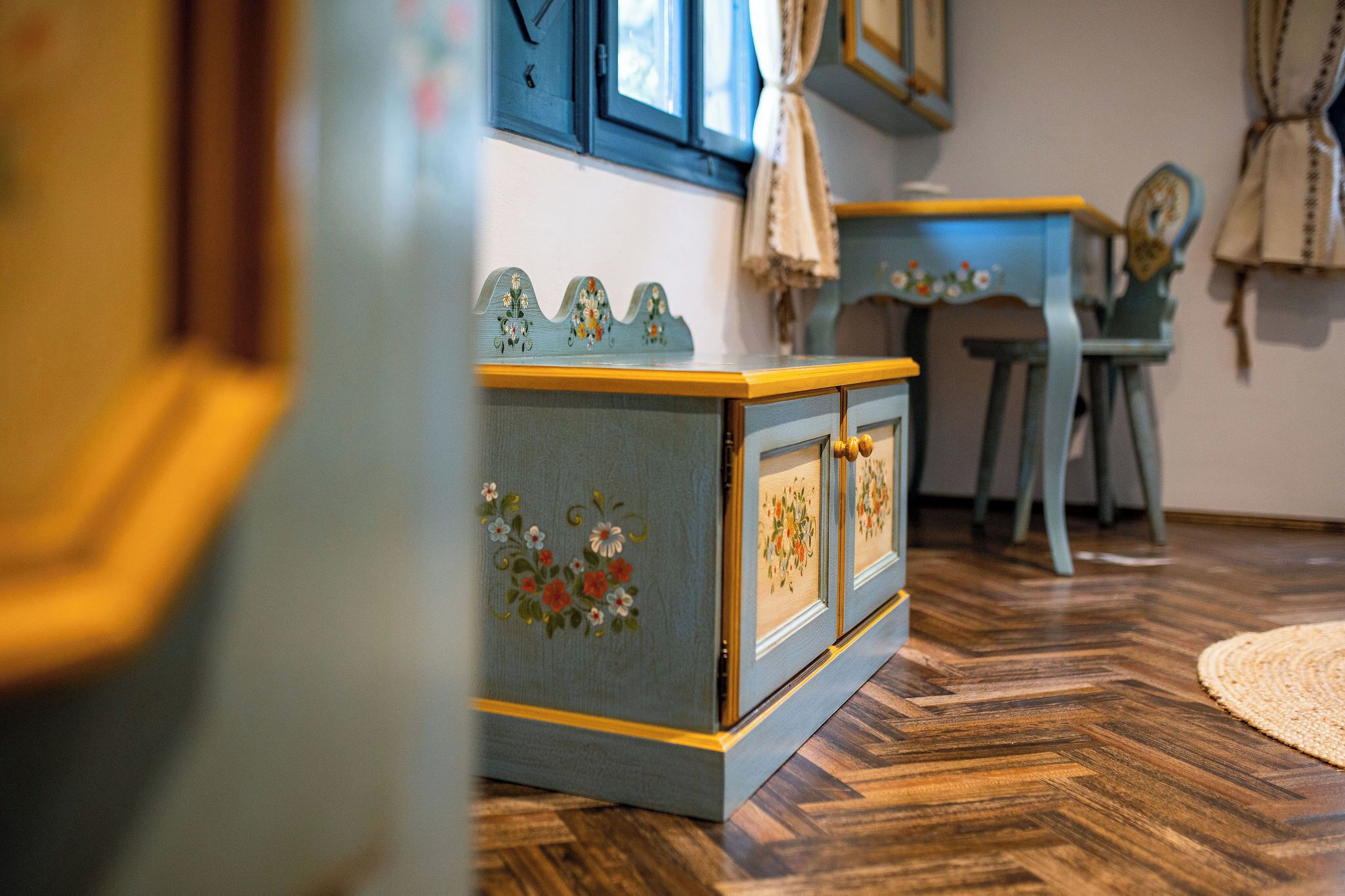 A room with a wooden floor, a blue cabinet with floral designs, a chair, and a table with a blue tablecloth.