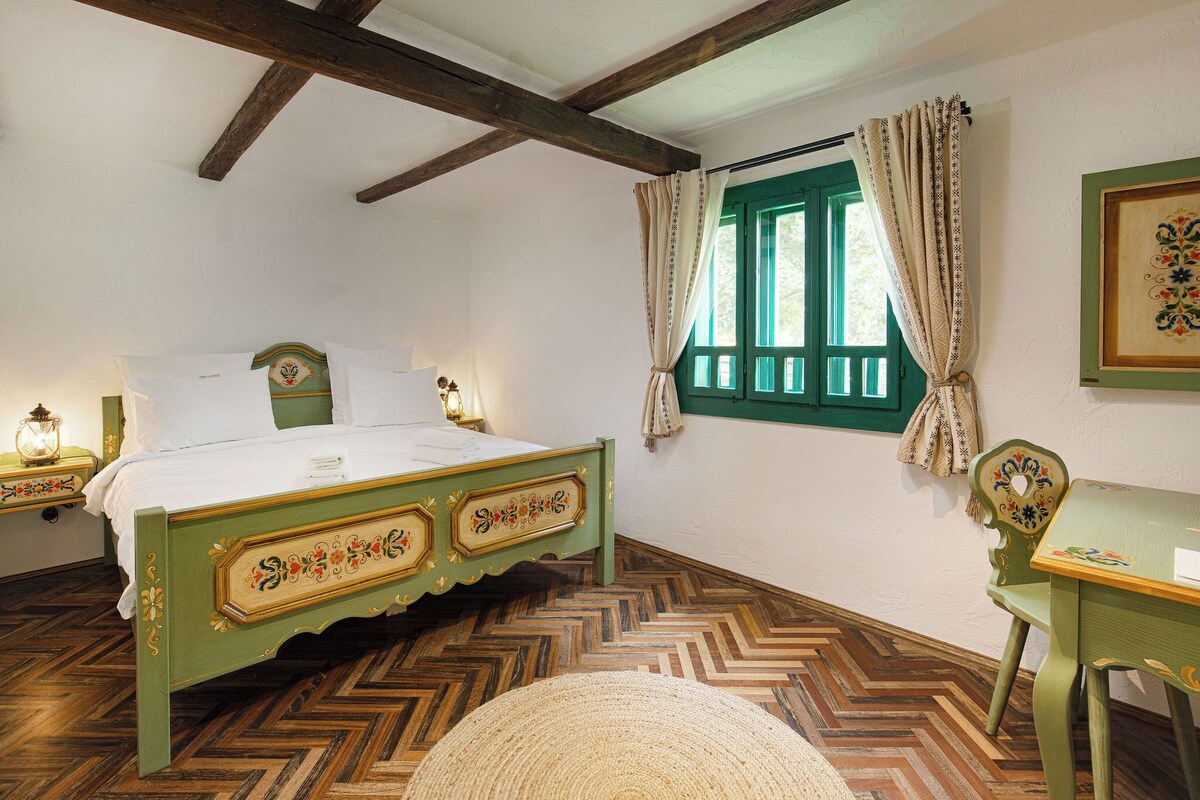 A traditional room with a wooden bed, a green chair, a desk, and a window with green shutters.