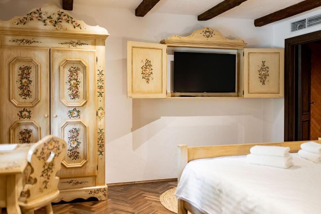 Quarto com cama, cadeira de madeira, guarda-roupa com estampas florais e televisão fixada sobre um armário.
