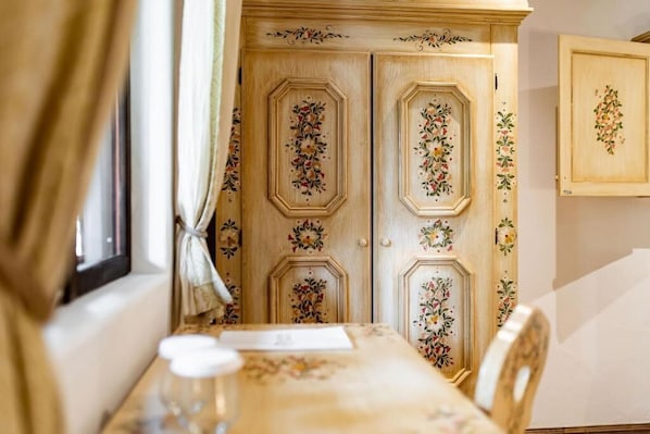 A room with a wooden door featuring floral carvings, a table with a floral tablecloth, and a window with curtains.