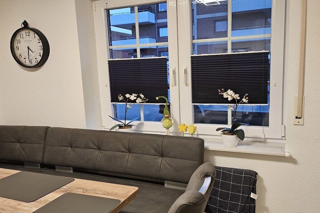 A living room with a sofa, a table, a clock, and a window with blinds and plants.