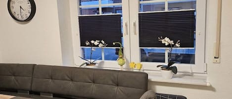 A living room with a sofa, a table, a clock, and a window with blinds and plants.
