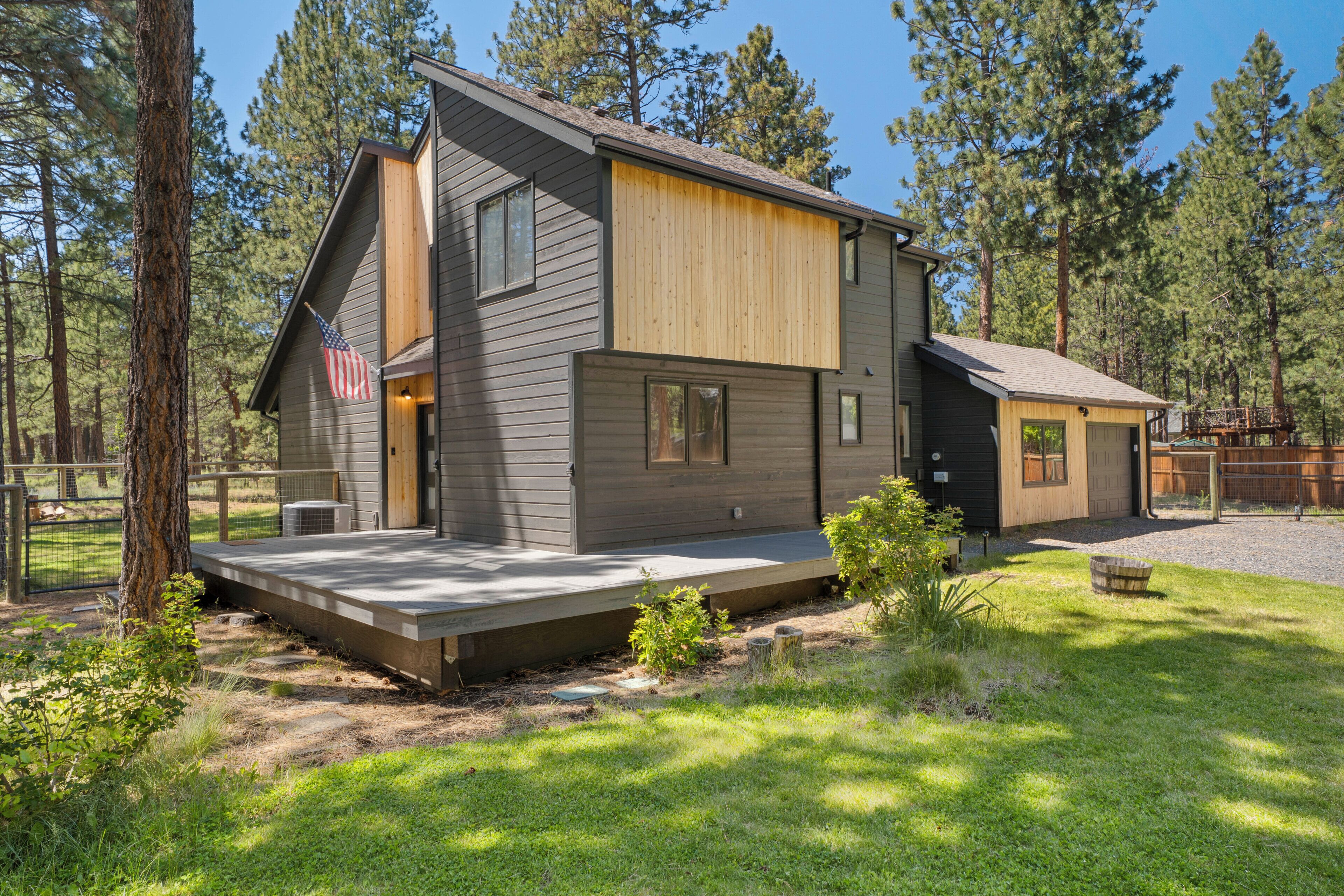 A modern two-story house with a wooden deck, a fenced yard, and a view of the surrounding forest.