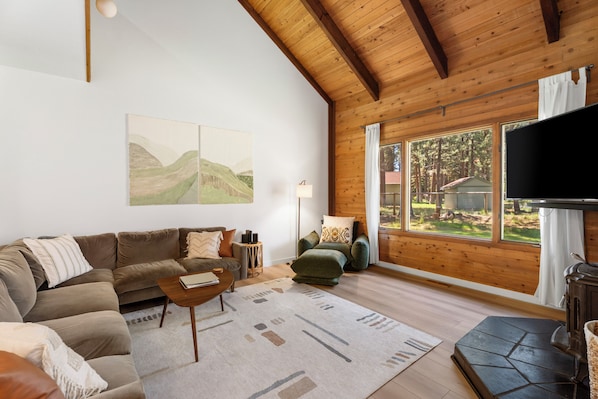 A cozy living room with a wooden ceiling, a large window with a view of the outdoors, a sofa, a coffee table, and a television.