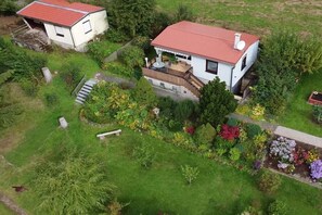 Une maison à deux étages, avec un toit rouge, entourée d’un jardin bien entretenu où poussent différentes plantes et fleurs.