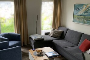 A living room with a grey sofa, a blue armchair, and a coffee table with books and a remote control. - Holiday home in Wangerland with sauna (Wangerland)