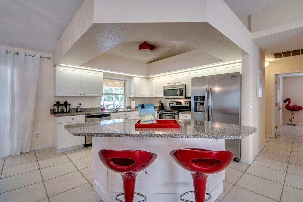Een moderne keuken met witte kasten, een centraal eiland met rode barstoelen, roestvrijstalen apparatuur en een rode plafondlamp.