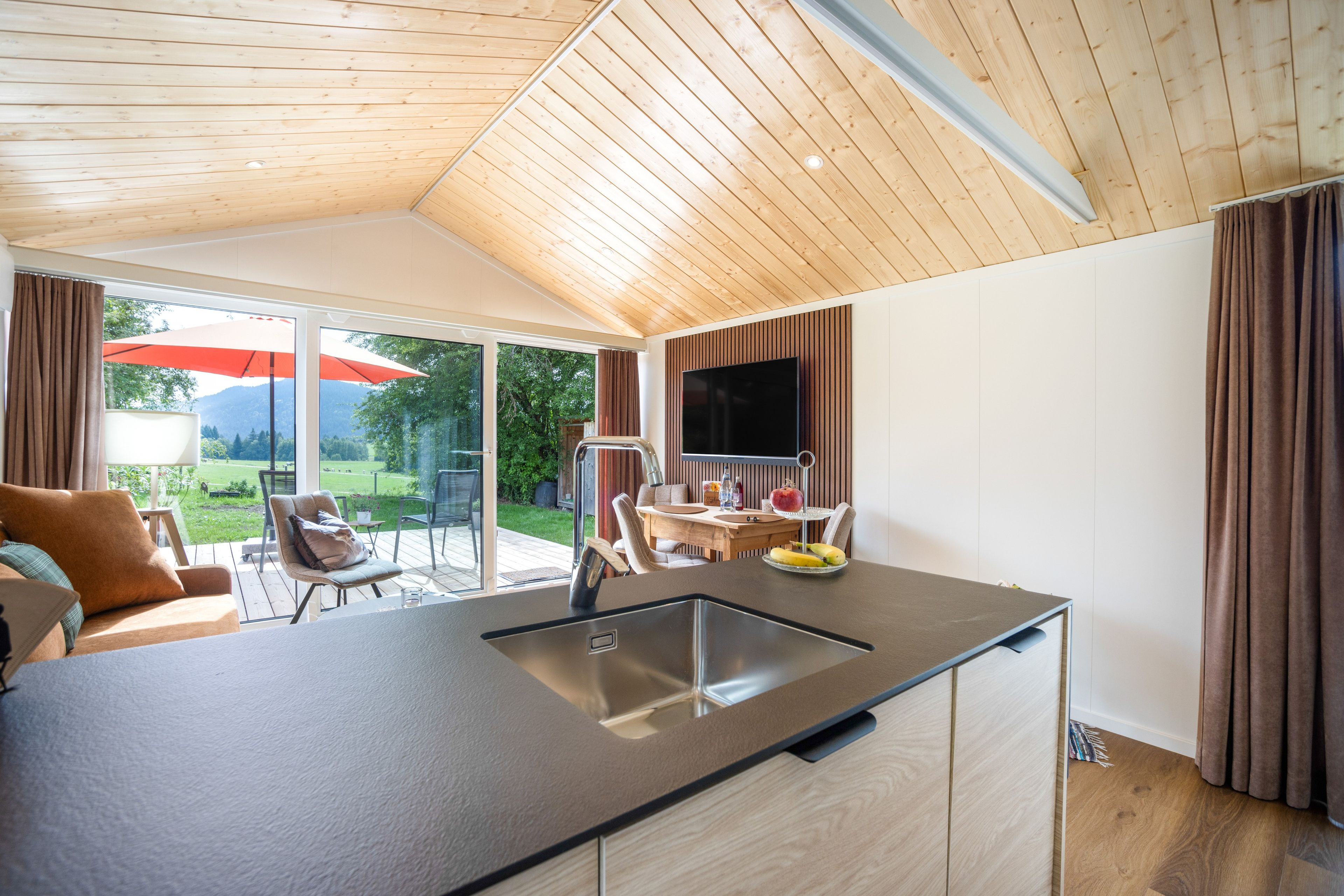 A modern kitchen with a large island, a dining area with outdoor seating, and a TV mounted on the wall.