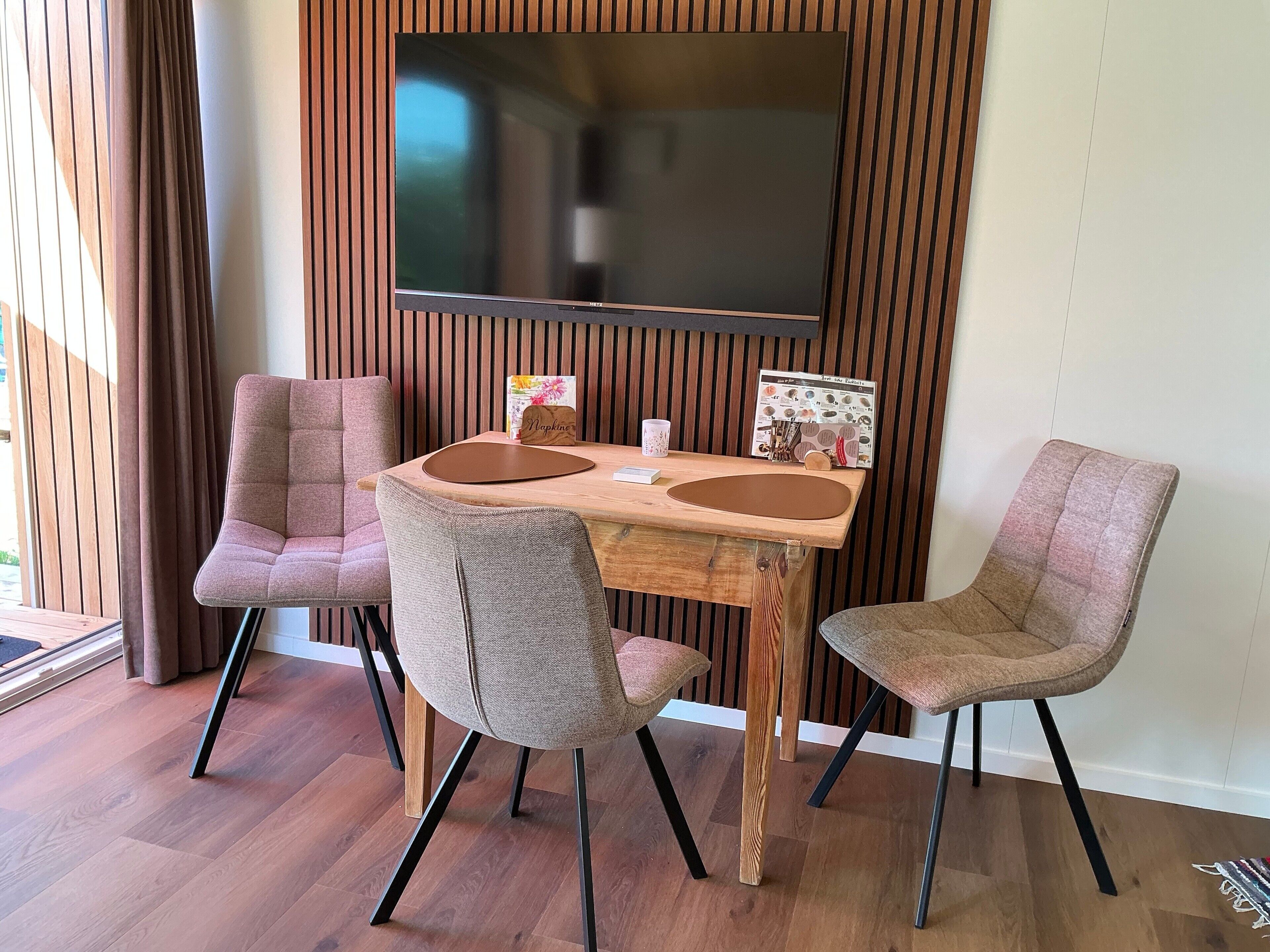 A dining area with a wooden table, four chairs, a flat-screen TV mounted on a wooden wall, and a window with curtains.