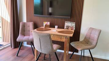 A dining area with a wooden table, four chairs, a flat-screen TV mounted on a wooden wall, and a window with curtains.