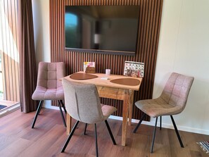 A dining area with a wooden table, four chairs, a flat-screen TV mounted on a wooden wall, and a window with curtains. - Tiny Home "Auszeit" with Mountain View, Wi-Fi, and Air Conditioning (Scheidegg)