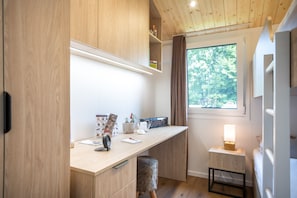 Ein kompakter, moderner Raum mit Schreibtisch aus Holz, einer kleinen Hocker, einem Fenster mit Blick auf Grünflächen und einem Nachttisch mit Lampe.