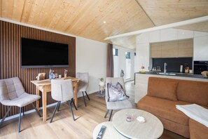 A modern living room with a flat-screen TV, a sofa, a dining area with chairs, and a kitchen in the background.