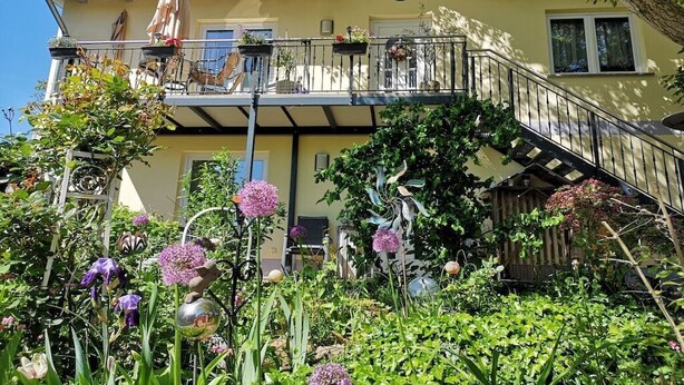 Ein zweistöckiges Haus mit Balkon, Treppe und einem Garten mit verschiedenen Pflanzen und Blumen.