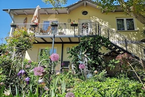 Ein zweistöckiges Haus mit Balkon, Treppe und einem Garten mit verschiedenen Pflanzen und Blumen.