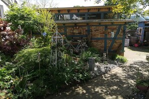 Een gebouw van baksteen met een houten dak, omringd door weelderige begroeiing en een geplaveid pad.
