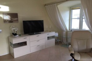 Ein modernes Wohnzimmer mit einem Flachbildfernseher auf einer weißen Kommode, einem Glastisch, einem beigen Ledersessel und einem Fenster mit Vorhängen.