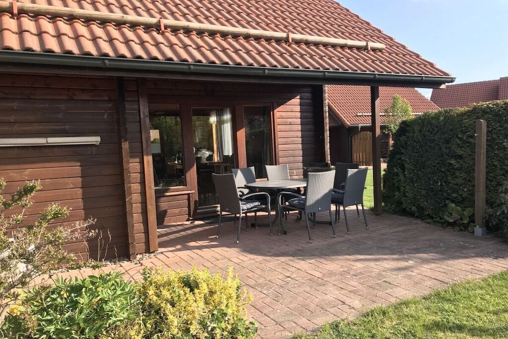 Een houten gebouw met een pannendak, een terras met een tafel en stoelen, en een haag op de achtergrond.