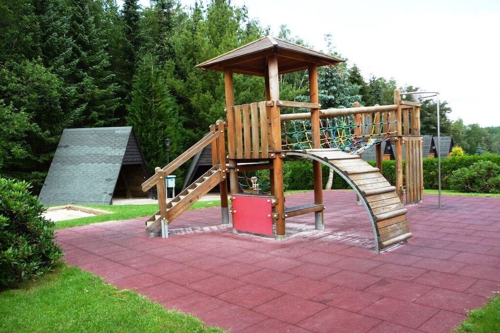 A wooden playground structure with a slide and climbing elements, surrounded by a paved area and greenery.