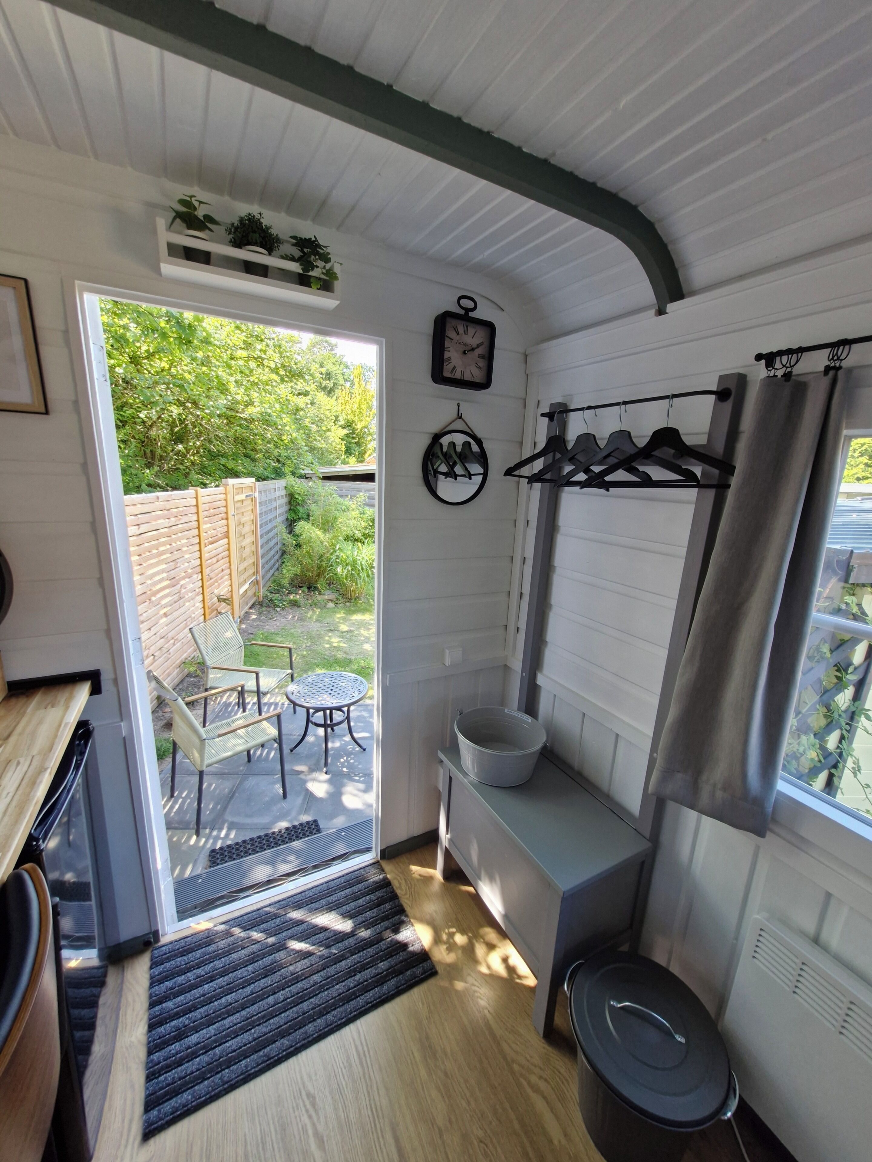 A compact room with a wooden bench, a small table, a hanging clothes rack, and a window with curtains.