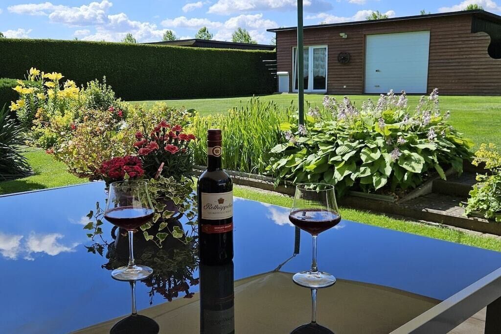 A table with two wine glasses and a bottle of wine, set outdoors with a view of a house and garden.