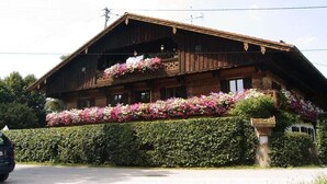 Una cabaña de madera con balcón decorado con flores, un seto en la parte delantera y cielo despejado.