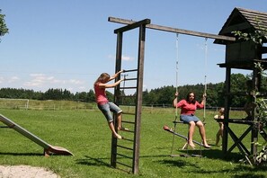 Puinen keinuteline, jossa on tikkaat, liuku ja pieni puinen rakennelma ruohikkoisella niityllä.