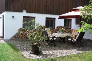 A patio area with a table and chairs, a potted plant, and a stack of firewood against a building with a white exterior and wooden siding.