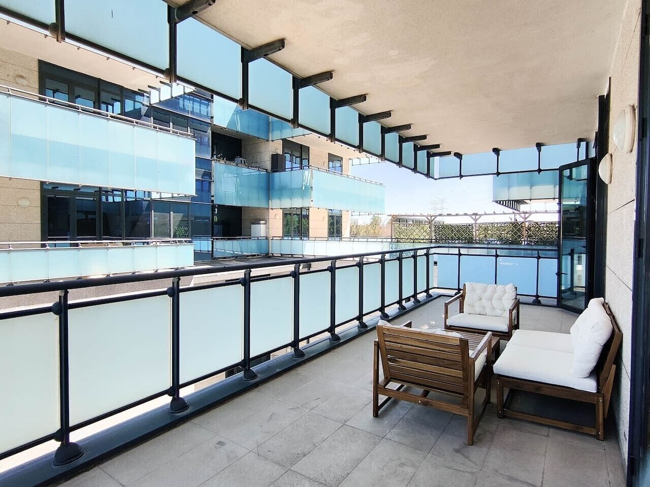 Un balcón con vista a un edificio moderno, amoblado con una mesa de madera y dos sillas blancas con cojines.