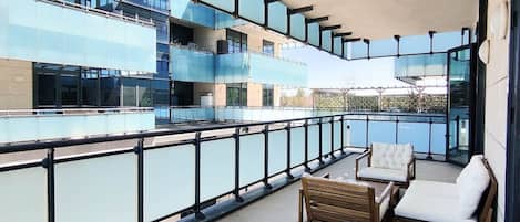A balcony with a view of a modern building, furnished with a wooden table and two white cushioned chairs.