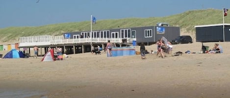 Un edificio frente a la playa con una terraza, una carpa azul y gente en la arena.